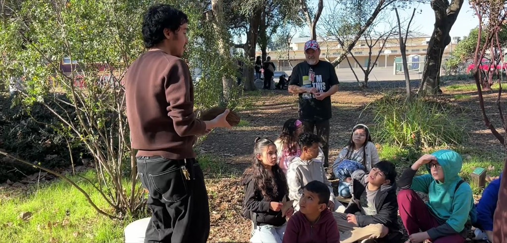Students in a classroom hold bare-root fruit trees in nursery pots alongside their worksheets